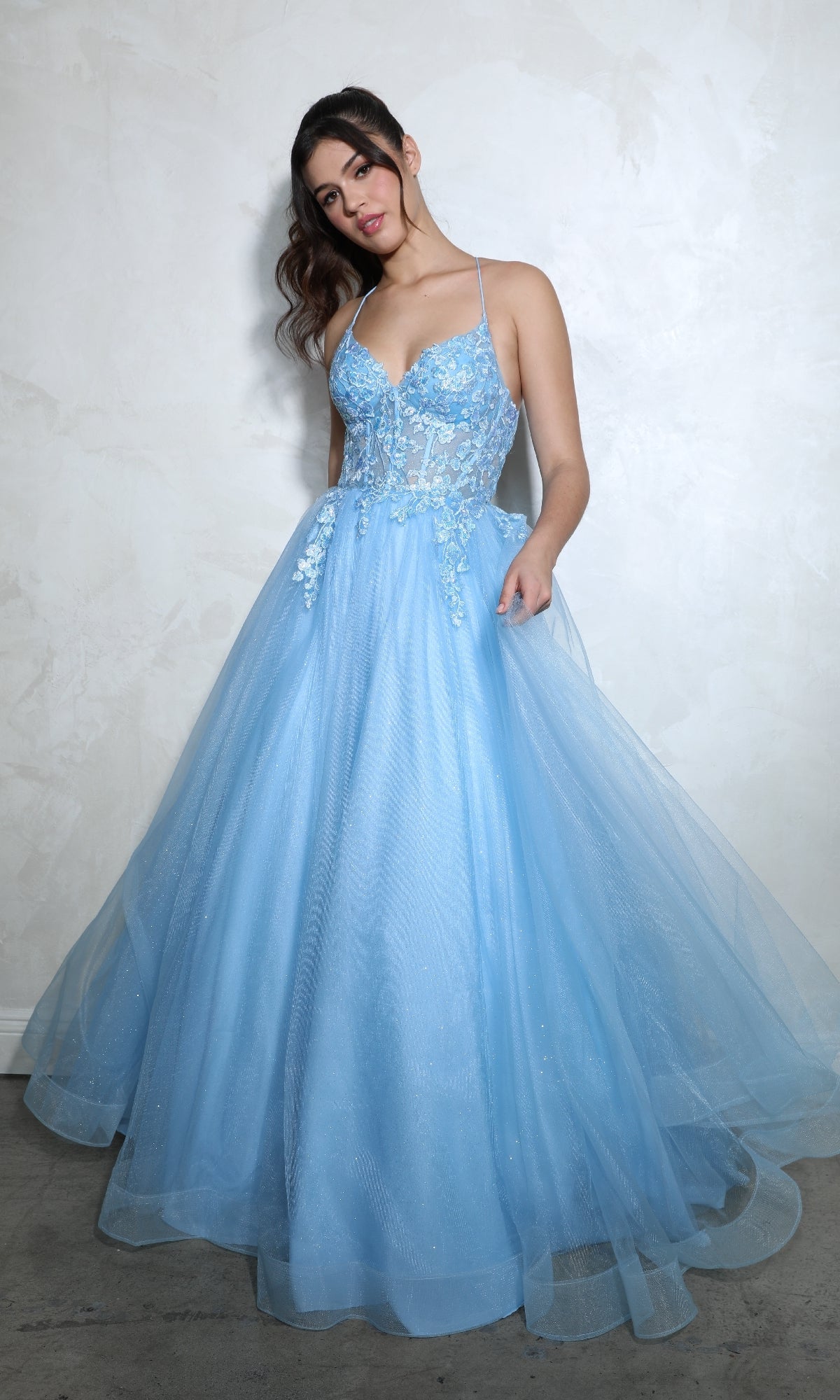 A woman poses against a light-colored textured wall in the Eureka 9960 Lace-Up Corset Long Prom Ball Gown, featuring a light blue, floor-length design with floral lace details and thin straps.